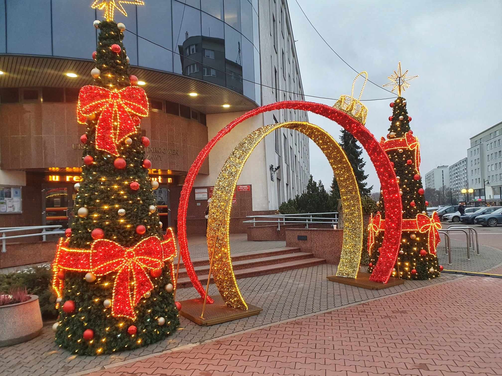 Ozdobione, oświetlone choinki i łuk z lampek przed budynkiem z przeszkloną fasadą, dekoracje świąteczne.