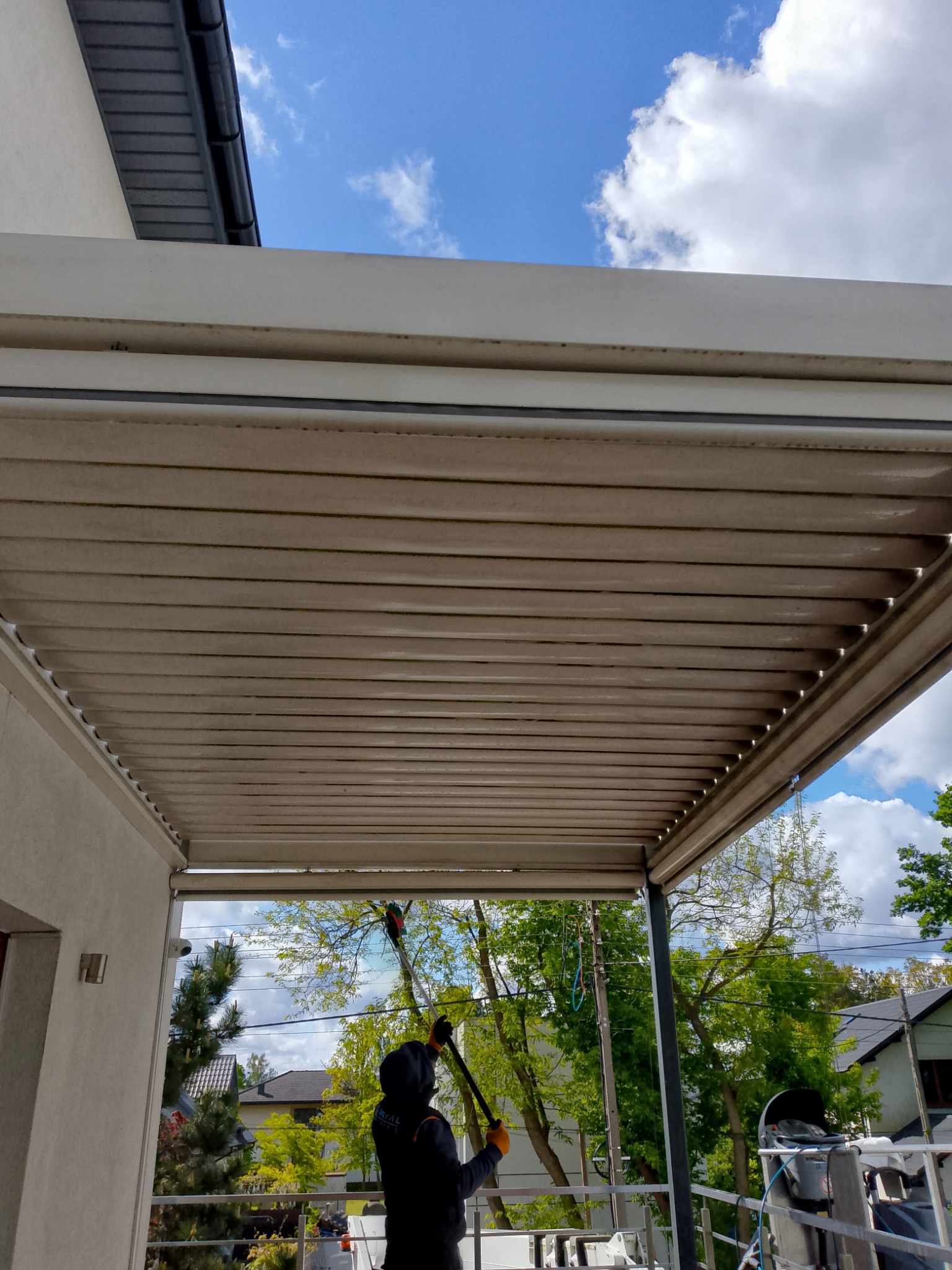 Osoba w kombinezonie czyści zadaszenie tarasu lancą teleskopową. Widoczne zabrudzenia na konstrukcji i błękitne niebo w tle.