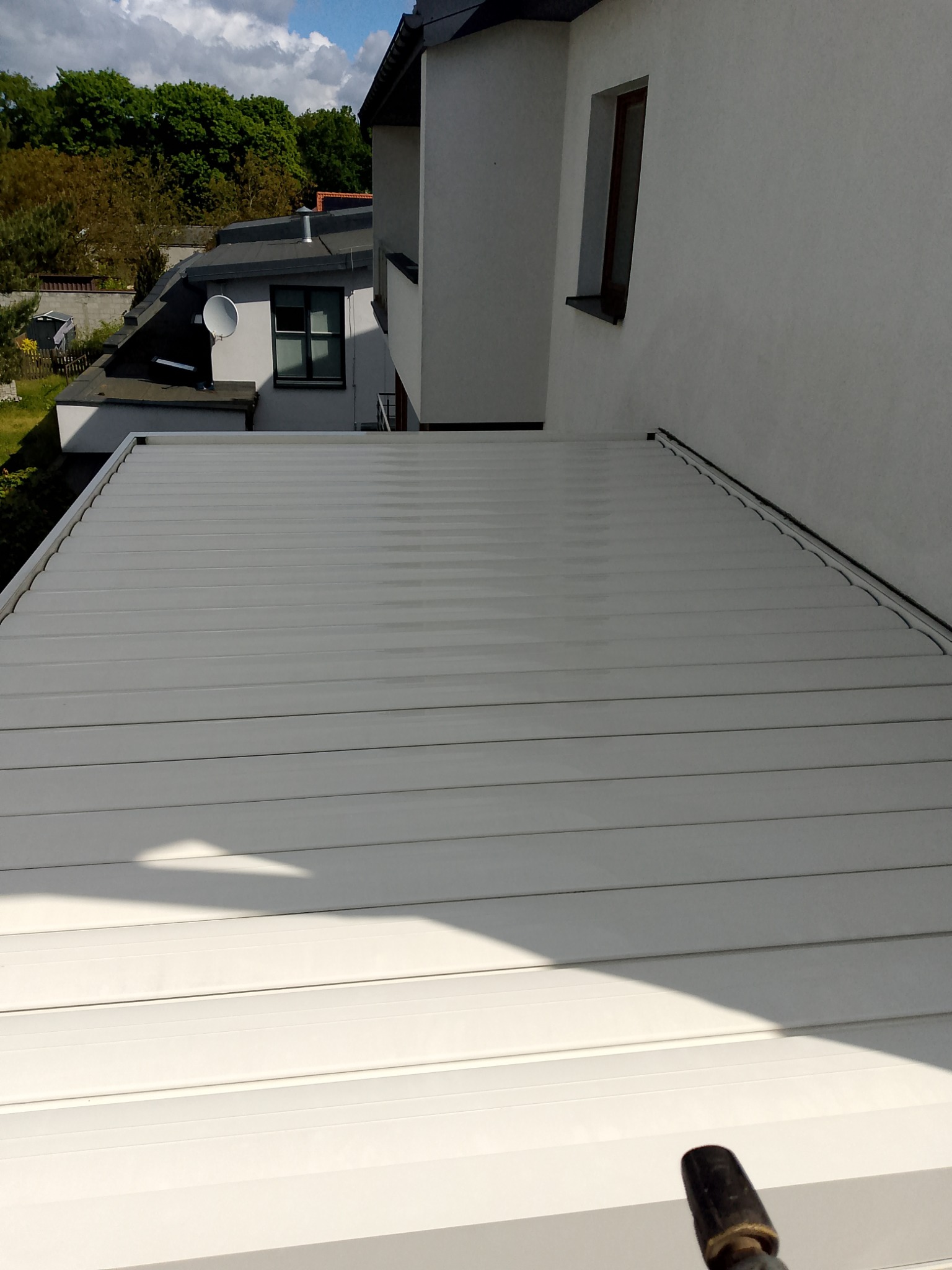 Biała, pozioma elewacja budynku z widocznymi liniami paneli, fragment budynku w tle, niebo z chmurami w oddali. Ujęcie z dołu.