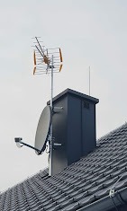 Antena telewizyjna Yagi-Uda i antena satelitarna zamontowane na kominie pokrytym grafitową blachą na dachu z ciemnoszarej dachówki ceramicznej.