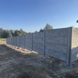 Twoje Ogrodzenie 24 - Długi, szary, betonowy płot o fakturze kamienia, z widocznymi słupkami i fundamentami, ciągnący się wzdłuż pola w słoneczny dzień.