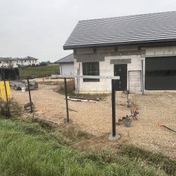 Twoje Ogrodzenie 24 - Wkopane słupki z poziomnicą w trakcie montażu ogrodzenia na posesji z domem w stanie surowym, widoczne narzędzia i materiały budowlane.