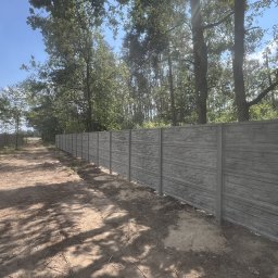 Twoje Ogrodzenie 24 - Długi, szary mur z prefabrykowanych elementów imitujących kamień, oddzielający teren zalesiony, widoczny w perspektywie wzdłuż polnej drogi.