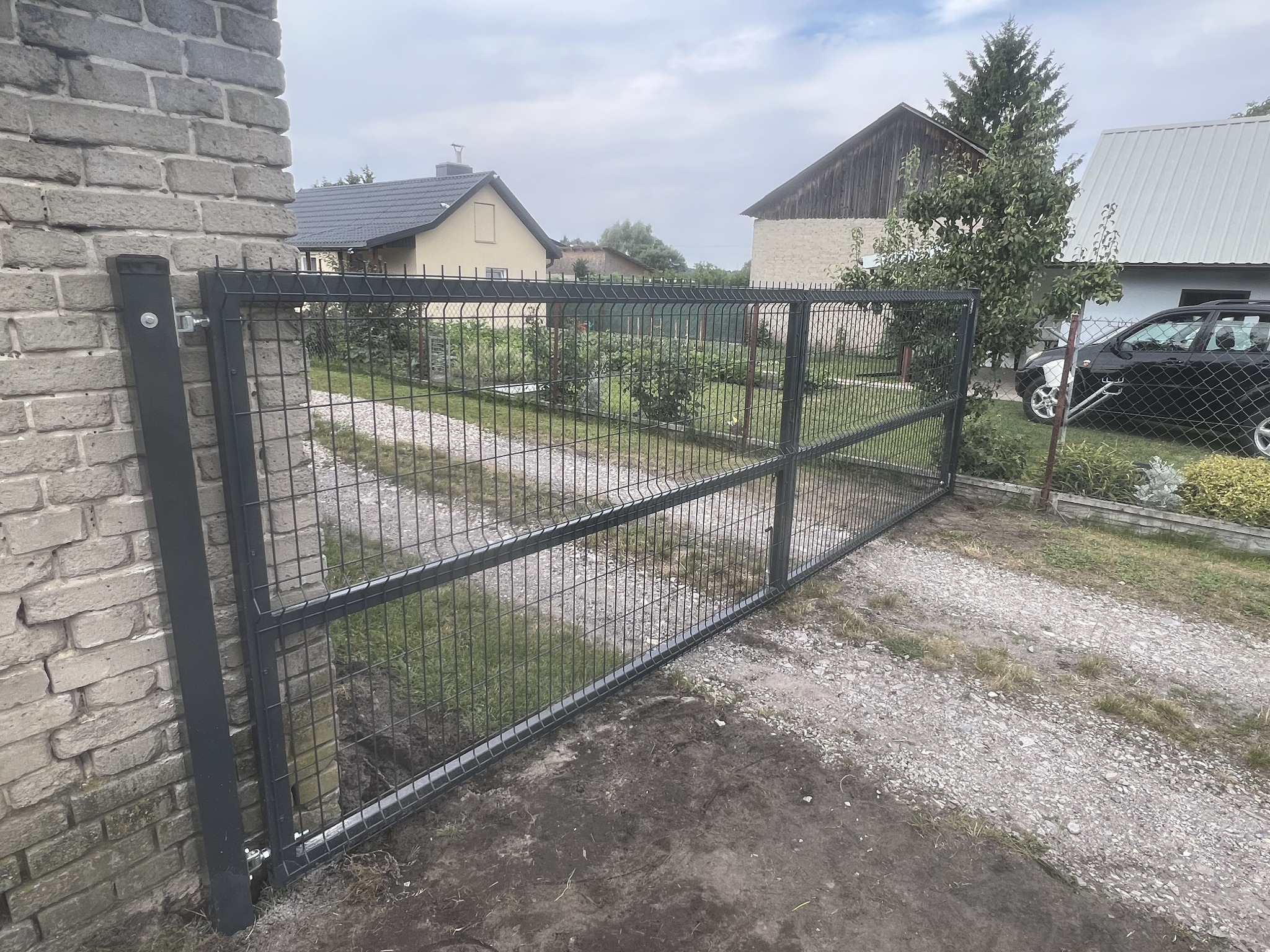 Otwarta, nowoczesna brama z paneli metalowych w kolorze grafitowym, zamontowana na murowanym słupku z cegły, prowadząca na żwirową drogę wjazdową; w tle widoczne budynki mieszkalne i zieleń.