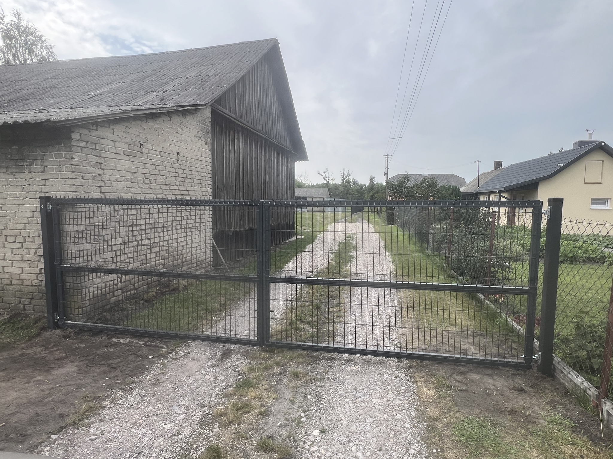 Nowoczesna, metalowa brama panelowa w kolorze grafitowym, z widocznym szarym budynkiem gospodarczym w tle i żwirową drogą prowadzącą w głąb posesji.