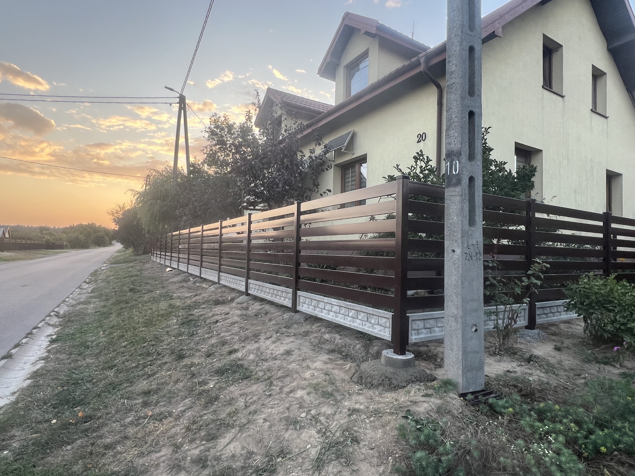 Nowoczesne ogrodzenie z poziomych, ciemnobrązowych paneli, z betonową podmurówką, w perspektywie ulicy i domu z numerem 20, oświetlone zachodzącym słońcem.