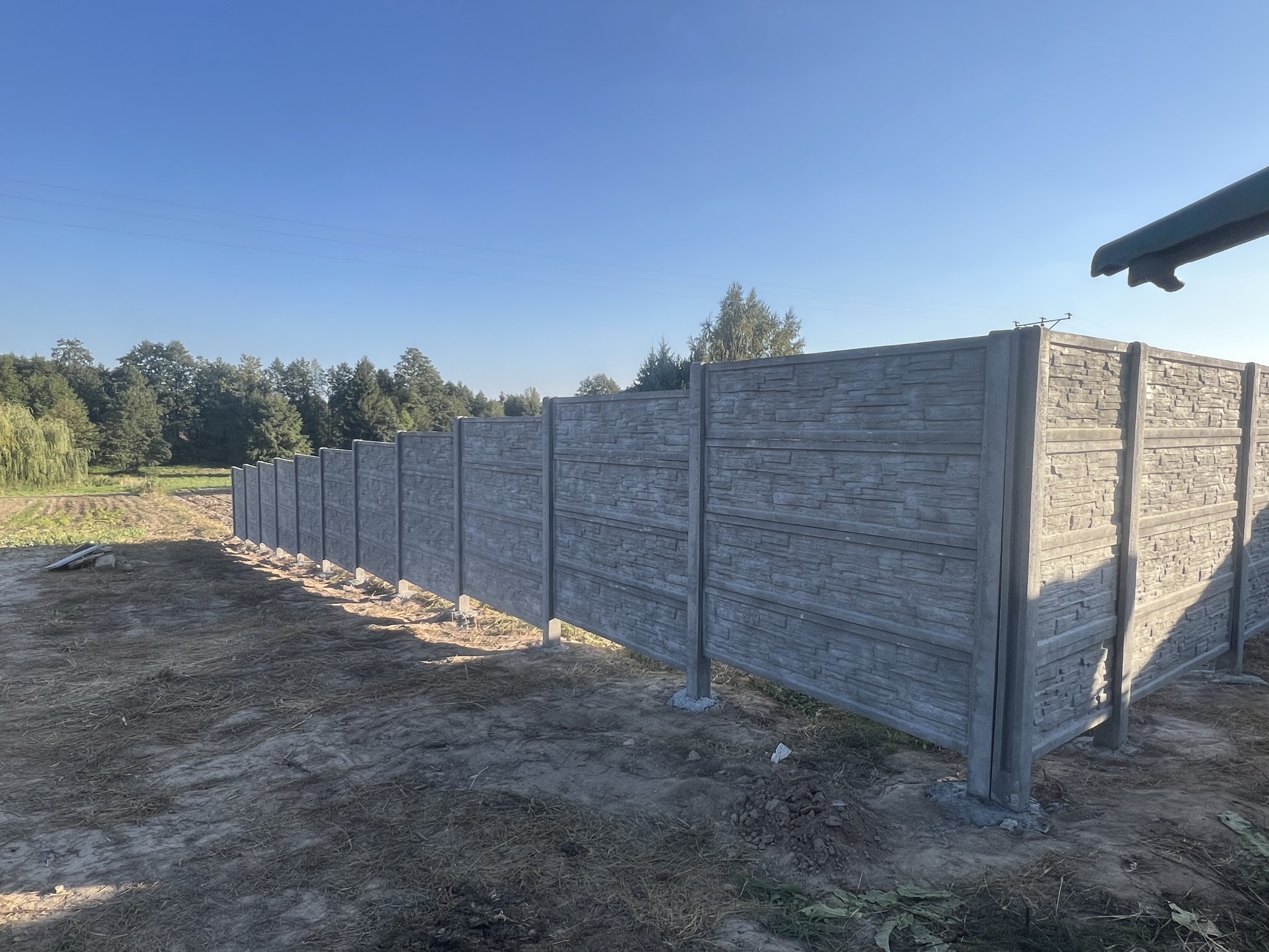 Długi, szary, betonowy płot o fakturze kamienia, z widocznymi słupkami i fundamentami, ciągnący się wzdłuż pola w słoneczny dzień.