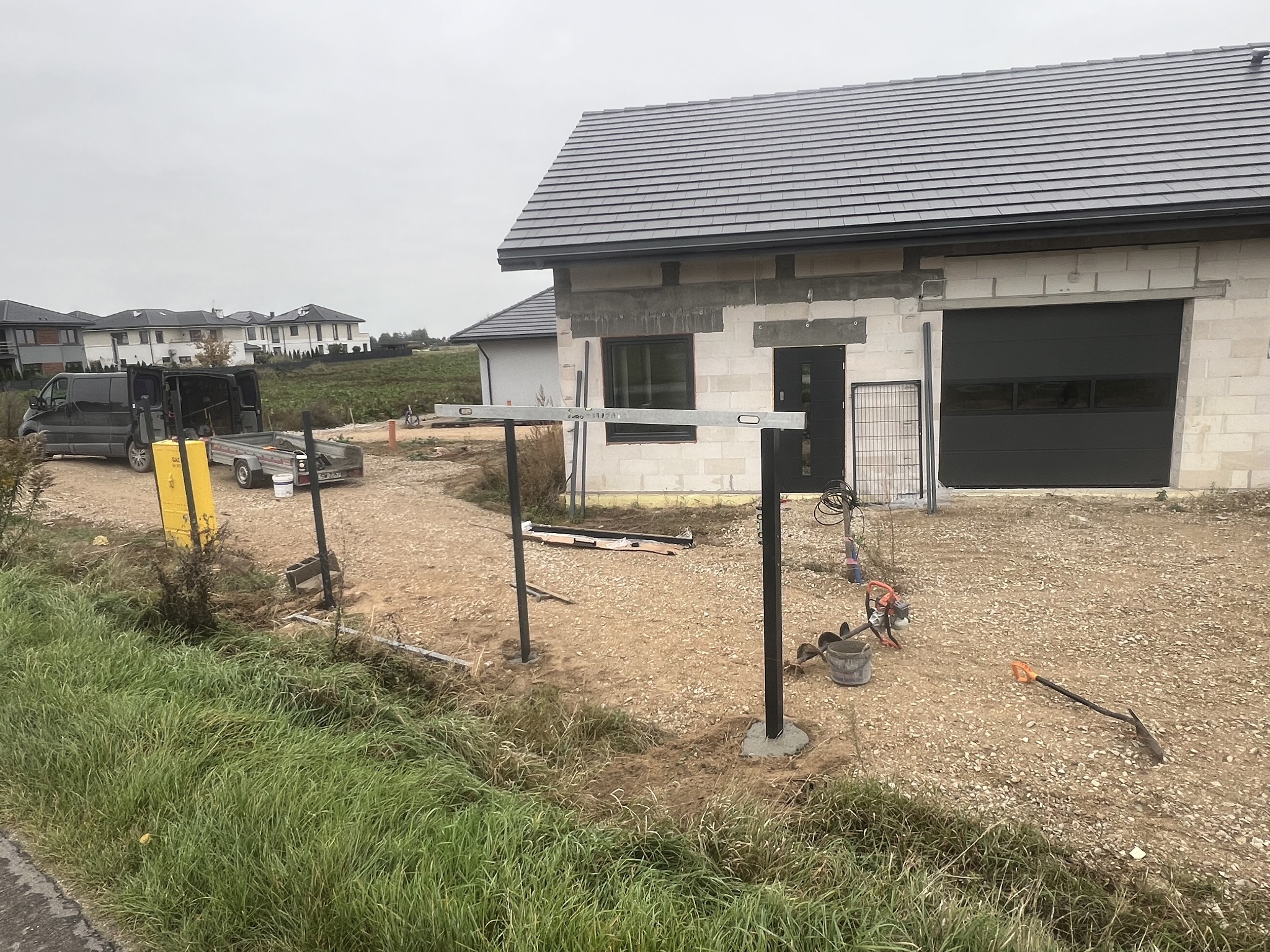 Wkopane słupki z poziomnicą w trakcie montażu ogrodzenia na posesji z domem w stanie surowym, widoczne narzędzia i materiały budowlane.