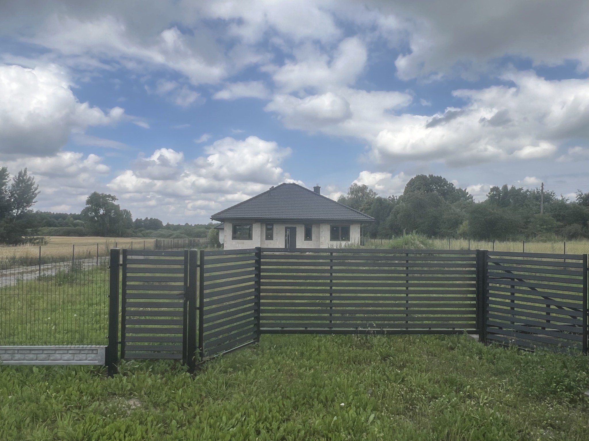 Nowoczesne, poziome, ciemnoszare przęsła ogrodzeniowe z widokiem na dom w tle, na tle błękitnego nieba z obłokami.