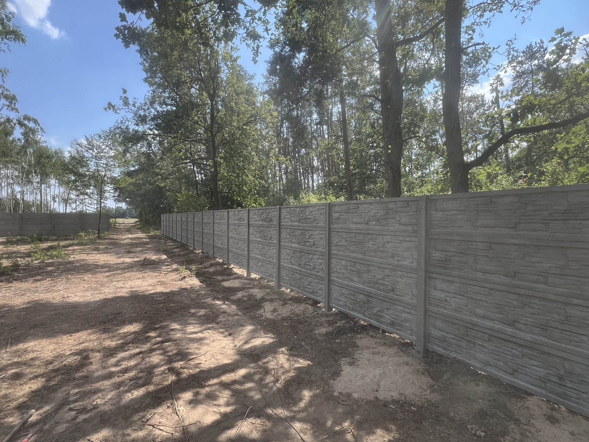 Długi, szary mur z prefabrykowanych elementów imitujących kamień, oddzielający teren zalesiony, widoczny w perspektywie wzdłuż polnej drogi.