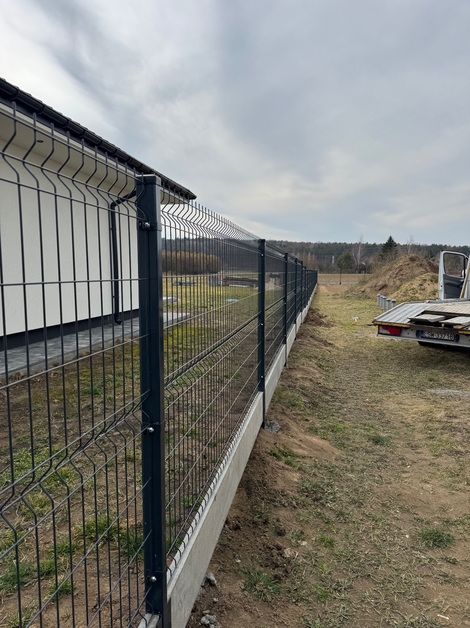 Długi, ciemny panelowy system ogrodzeniowy z podmurówką betonową, ciągnący się wzdłuż granicy posesji obok białego domu, z przyczepą i stertą ziemi w tle, pochmurne niebo.