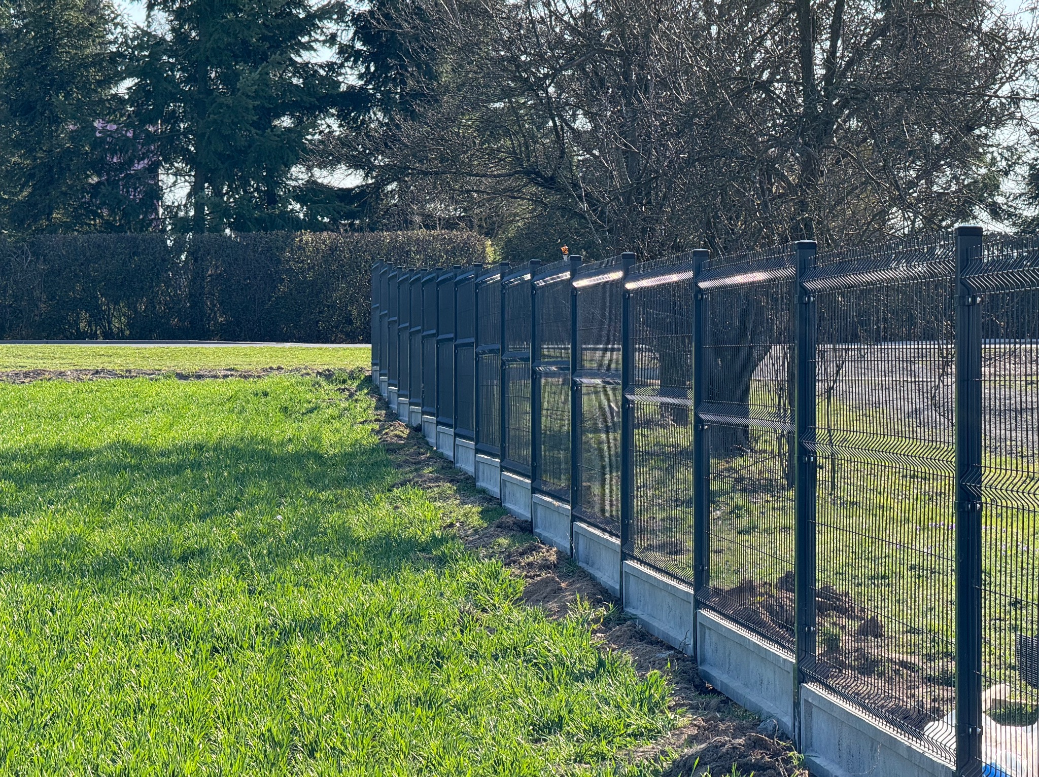 Długa, czarna, metalowa konstrukcja ogradzająca teren z betonową podmurówką, widoczna na tle zielonej trawy i drzew.