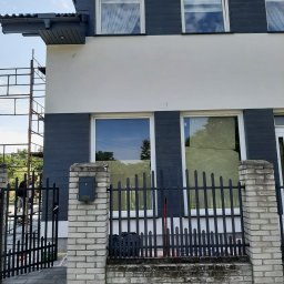 Wojciech Dudka - Dwukondygnacyjny dom z białą elewacją i ciemnoszarą okładziną drewnopodobną wokół okien, widoczne rusztowanie i pracownik, ogrodzenie z ceglanymi słupkami i metalowymi przęsłami, skrzynka pocztowa...