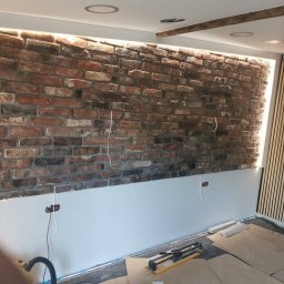 Wojciech Dudka - Ściana z cegły klinkierowej z podświetleniem LED w górnej części, dolna część ściany pomalowana na szaro, widoczne przewody elektryczne, listwy przypodłogowe i narzędzia na podłodze, panele ścienne...