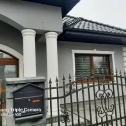 Elewacja domu w odcieniach szarości z białymi kolumnami i detalami. Widoczna brama z metaloplastyki i skrzynka pocztowa. Ujęcie z bliska, podkreślające estetykę wykończenia.