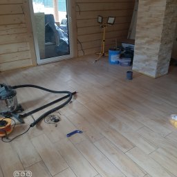 BudWojtus - Jasne wnętrze z nowo ułożoną podłogą z płytek drewnopodobnych, ścianami z drewnianych paneli i kolumną z kamienia dekoracyjnego. Widoczne narzędzia do prac wykończeniowych.