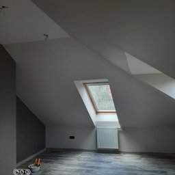 BudWojtus - Poddasze po remoncie z jasnymi ścianami i ciemnoszarą ścianą akcentową, skośnymi sufitami, oknem dachowym, grzejnikiem i panelami podłogowymi. Na podłodze narzędzia malarskie.