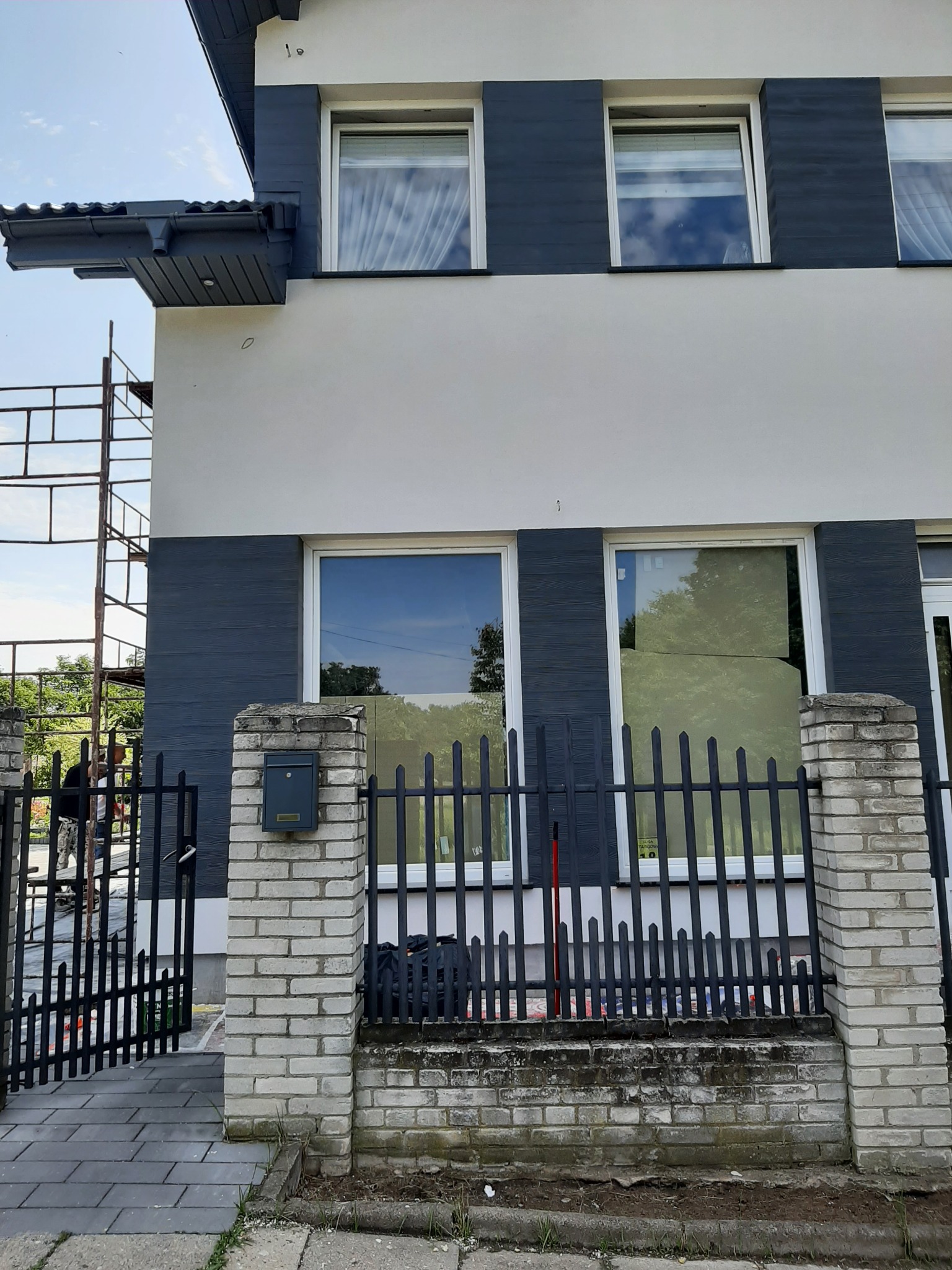 Dwukondygnacyjny dom z białą elewacją i ciemnoszarą okładziną drewnopodobną wokół okien, widoczne rusztowanie i pracownik, ogrodzenie z ceglanymi słupkami i metalowymi przęsłami, skrzynka pocztowa...