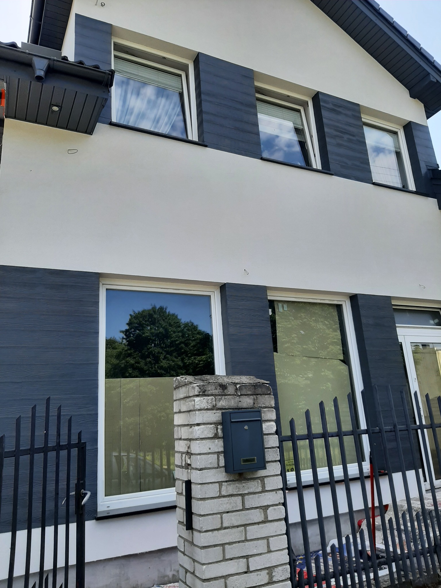 Dwukondygnacyjny dom z szarą elewacją imitującą drewno i białym tynkiem, widok zza czarnego ogrodzenia i murowanego słupka z szarą skrzynką pocztową.