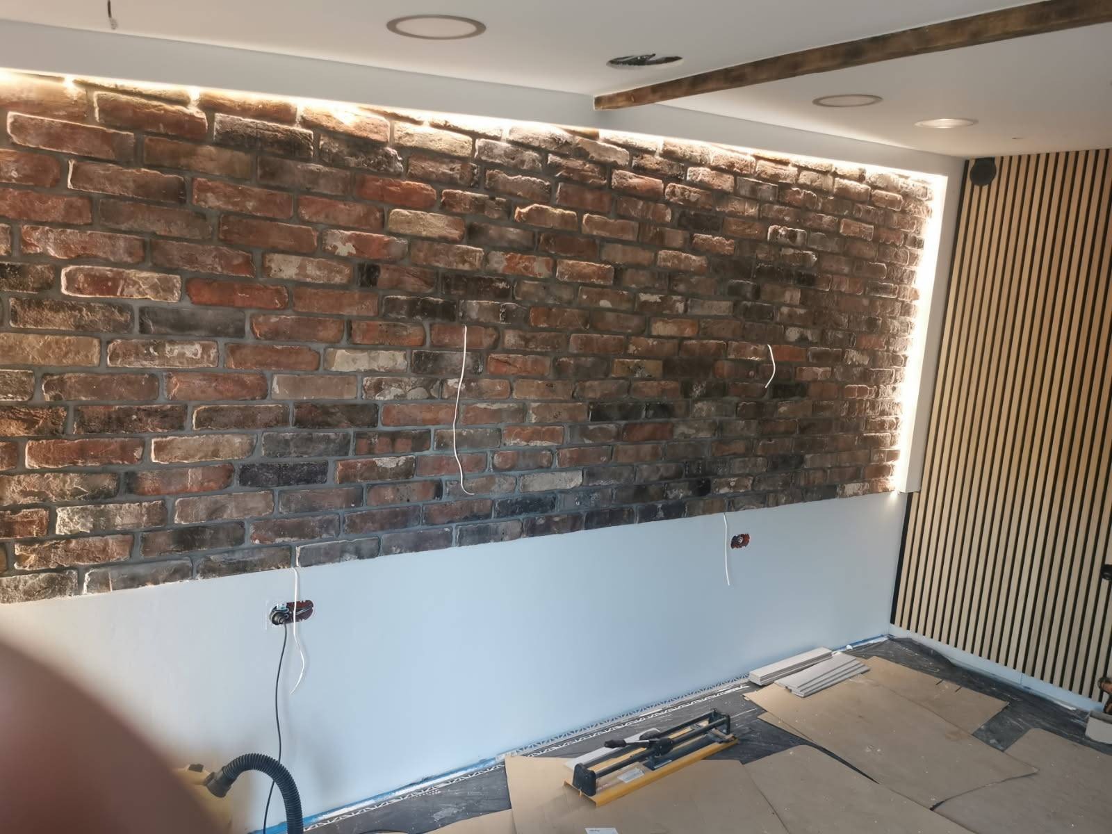 Ściana z cegły klinkierowej z podświetleniem LED w górnej części, dolna część ściany pomalowana na szaro, widoczne przewody elektryczne, listwy przypodłogowe i narzędzia na podłodze, panele ścienne...