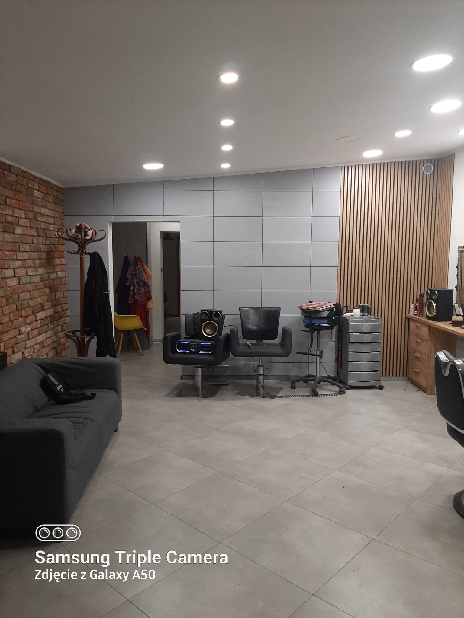Wnętrze salonu fryzjerskiego z szarą sofą, ceglaną ścianą, szarymi płytkami i trzema fotelami fryzjerskimi, jeden z głośnikiem na siedzeniu.