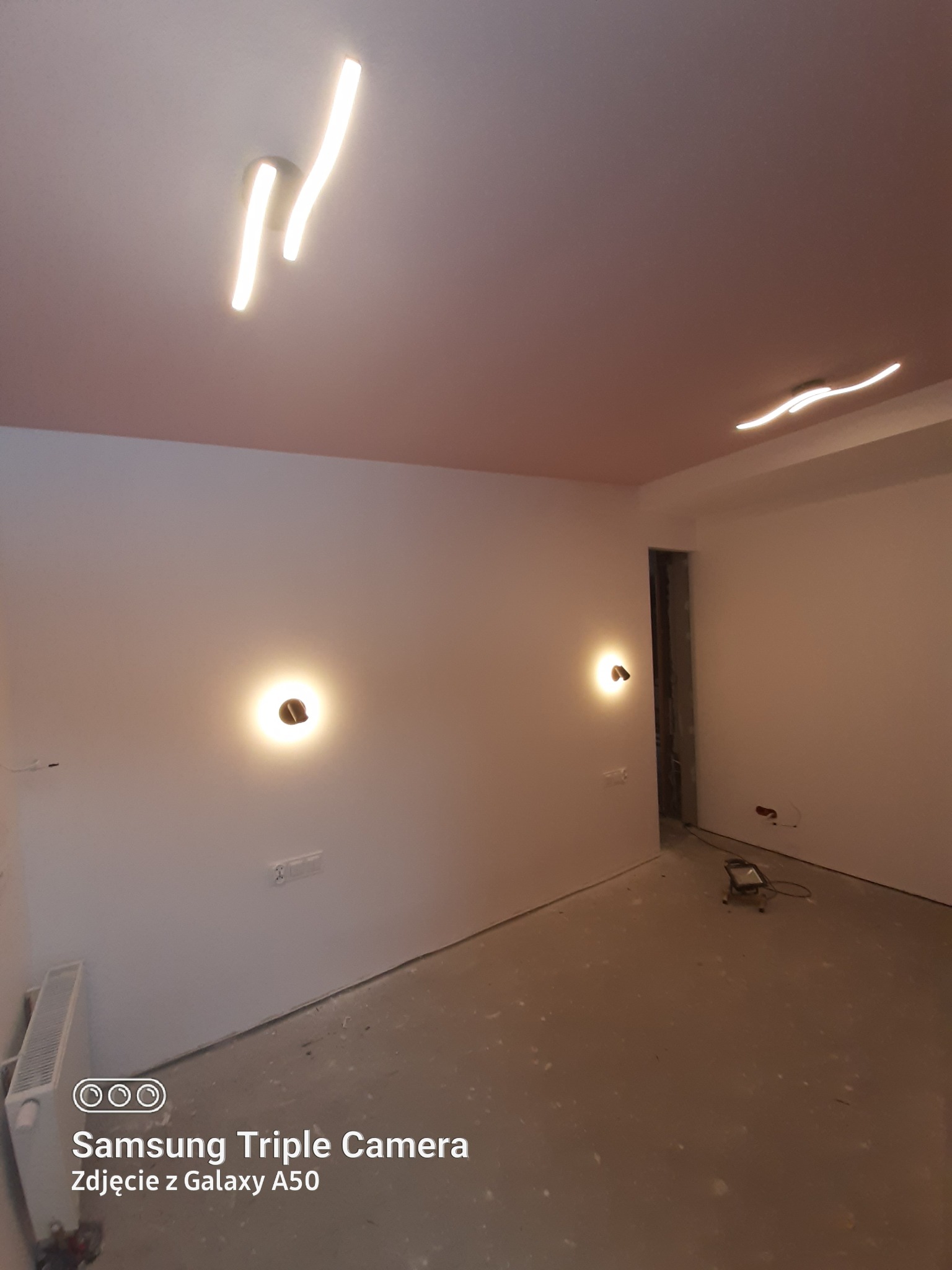 Wnętrze w trakcie prac wykończeniowych: pomalowany sufit na różowo, białe ściany z zamontowanymi lampami ściennymi, widoczny grzejnik i lampa podłogowa.