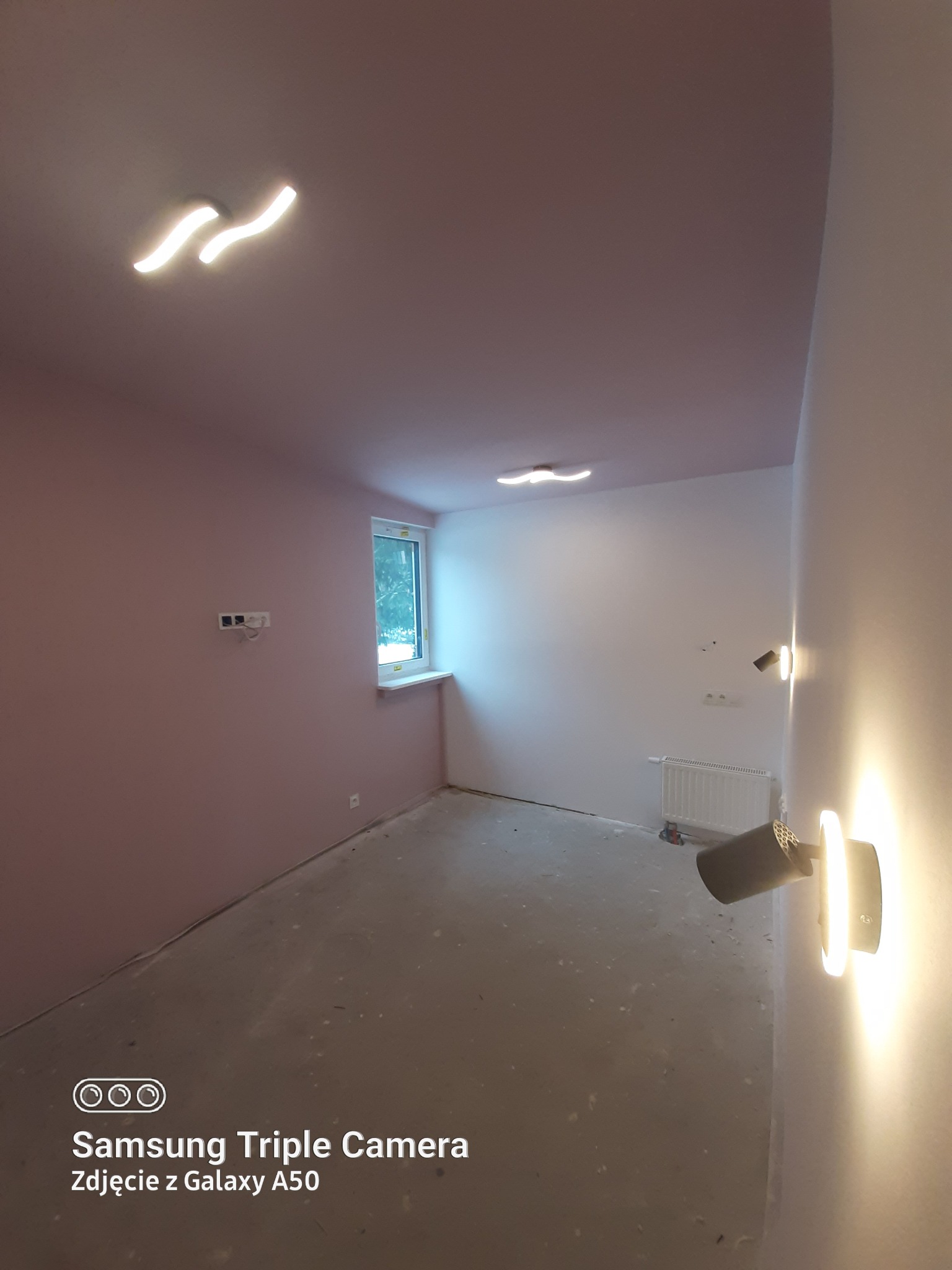 Pokój po malowaniu ścian na różowo i biało, z zamontowanymi lampami sufitowymi i ściennymi, widoczny grzejnik i okno.