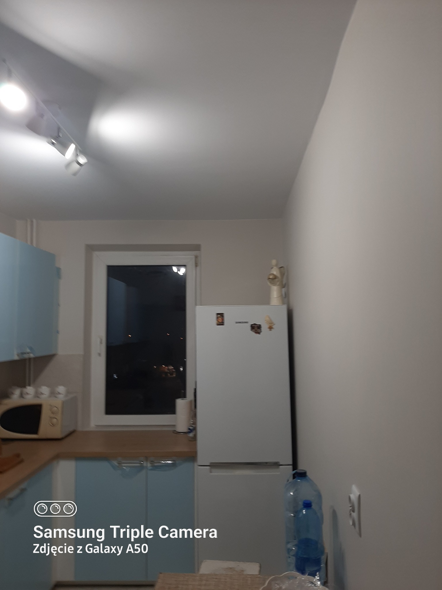 Kuchnia z niebieskimi szafkami, białym sprzętem AGD i widokiem na nocne miasto przez okno, uchwycona z perspektywy lekko z dołu.