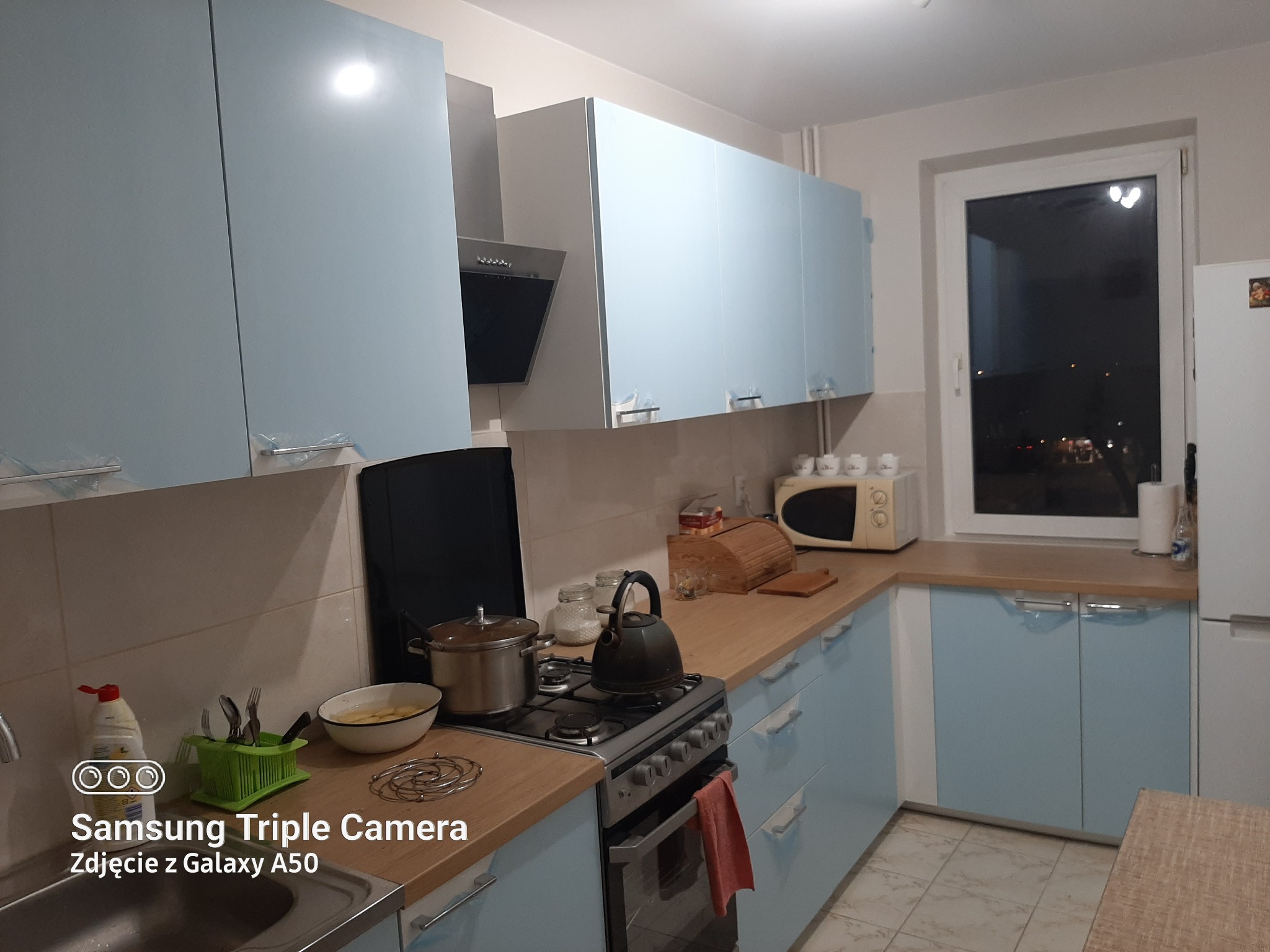 Jasna kuchnia z błękitnymi szafkami, czajnikiem na kuchence gazowej i widokiem nocnego miasta za oknem.