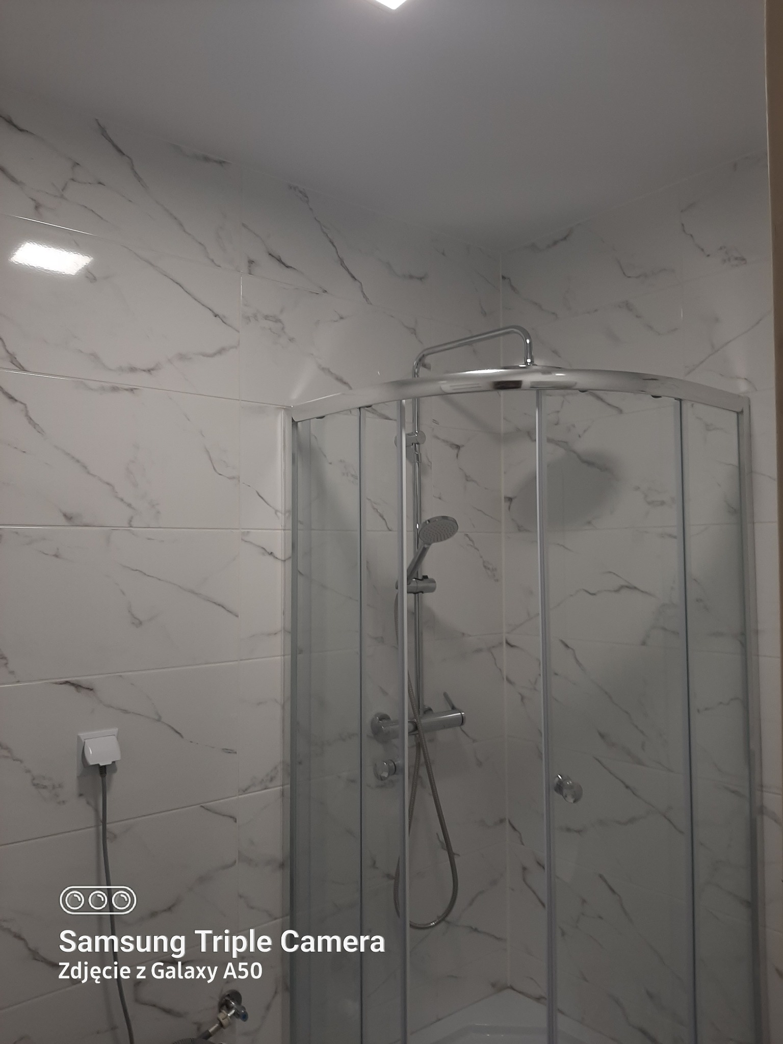 Narożna kabina prysznicowa z chromowanymi profilami i marmurkowymi płytkami na ścianach, widoczny biały kontakt elektryczny z kablem.