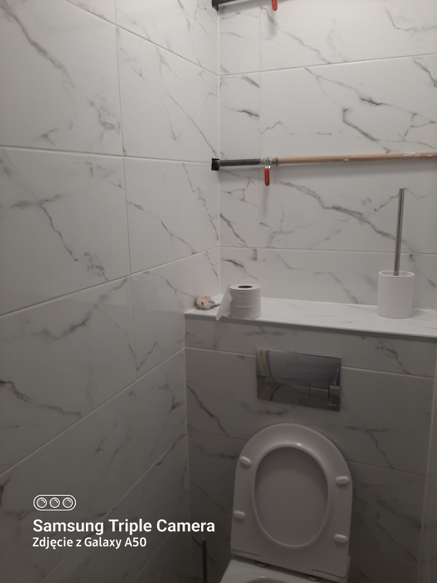 Biała łazienka z marmurowymi płytkami, widoczne rury z zaworami, toaleta i akcesoria łazienkowe.