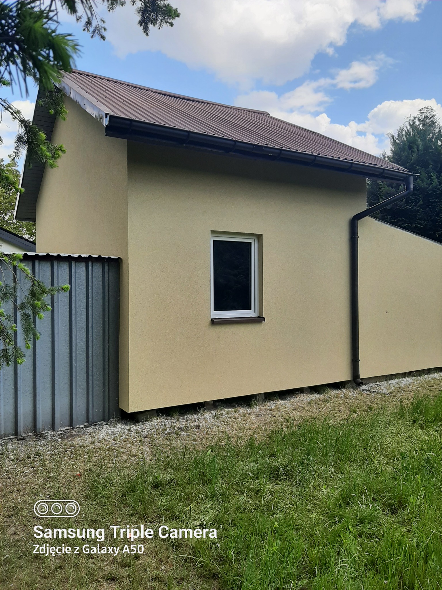 Otynkowana elewacja budynku z brązowym dachem i białym oknem, widoczna z poziomu trawnika w słoneczny dzień. Obok blaszane ogrodzenie.