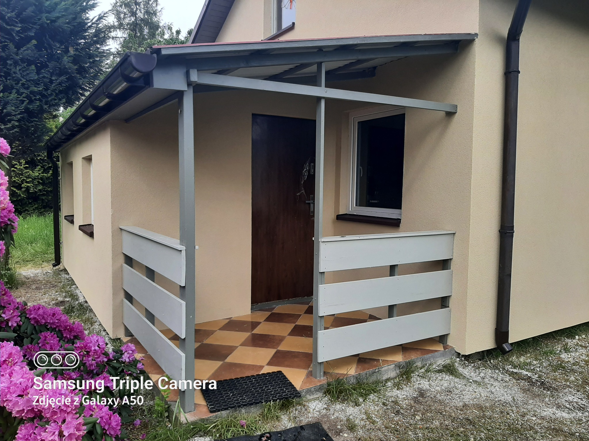 Wykończony, ocieplony dom z zadaszonym wejściem, ozdobiony kwiatami. Elewacja w odcieniach beżu, brązowe rynny, szara konstrukcja zadaszenia i biała balustrada.
