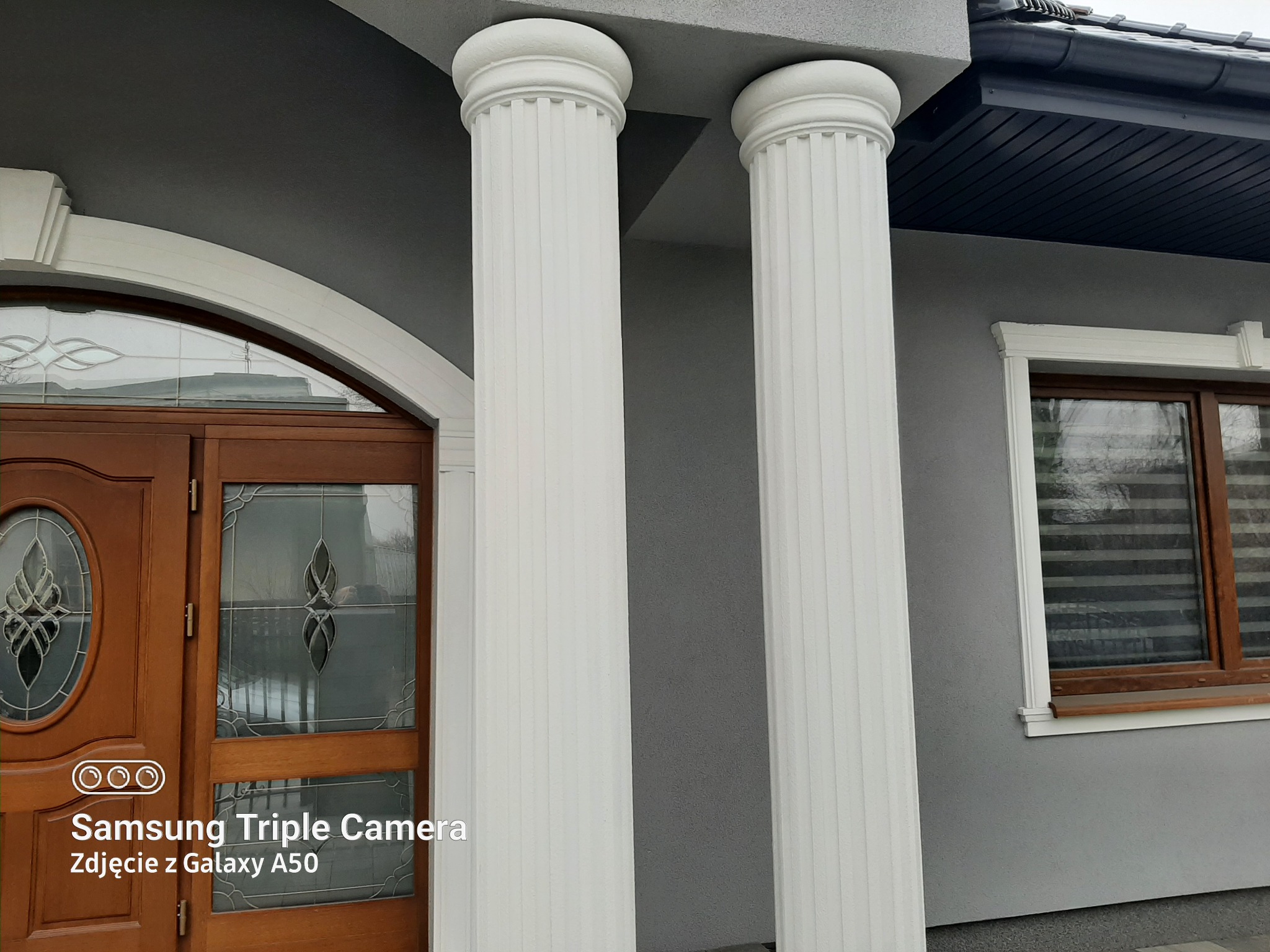 Szare wykończenie elewacji domu z dwiema białymi kolumnami, drewnianymi drzwiami z ozdobnym przeszkleniem i oknem z drewnianą ramą.