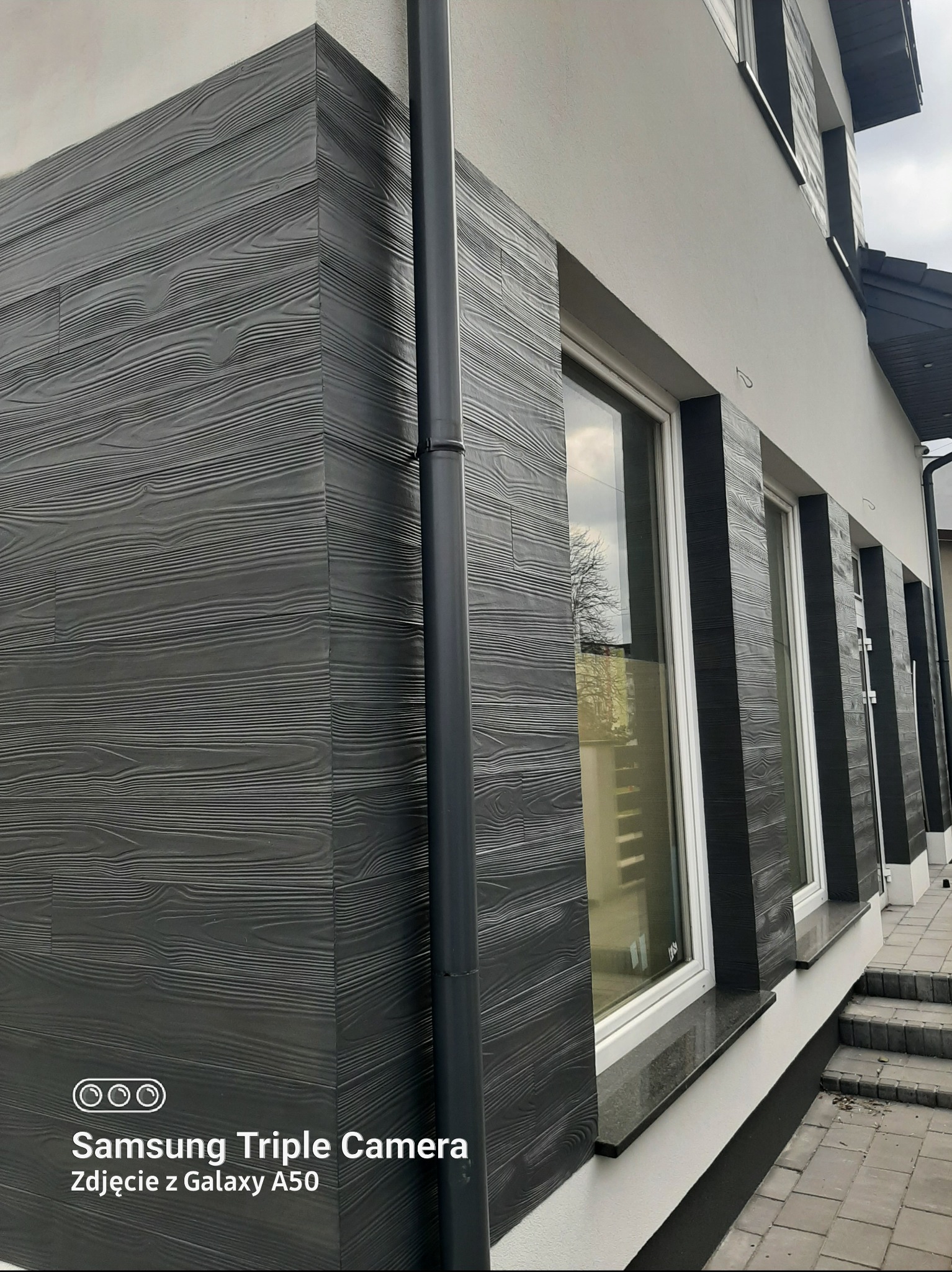 Elewacja domu z szarymi panelami imitującymi drewno, widoczne okna z białymi ramami i szarym parapetem, rynna. Ujęcie z dołu, podkreślające strukturę elewacji.