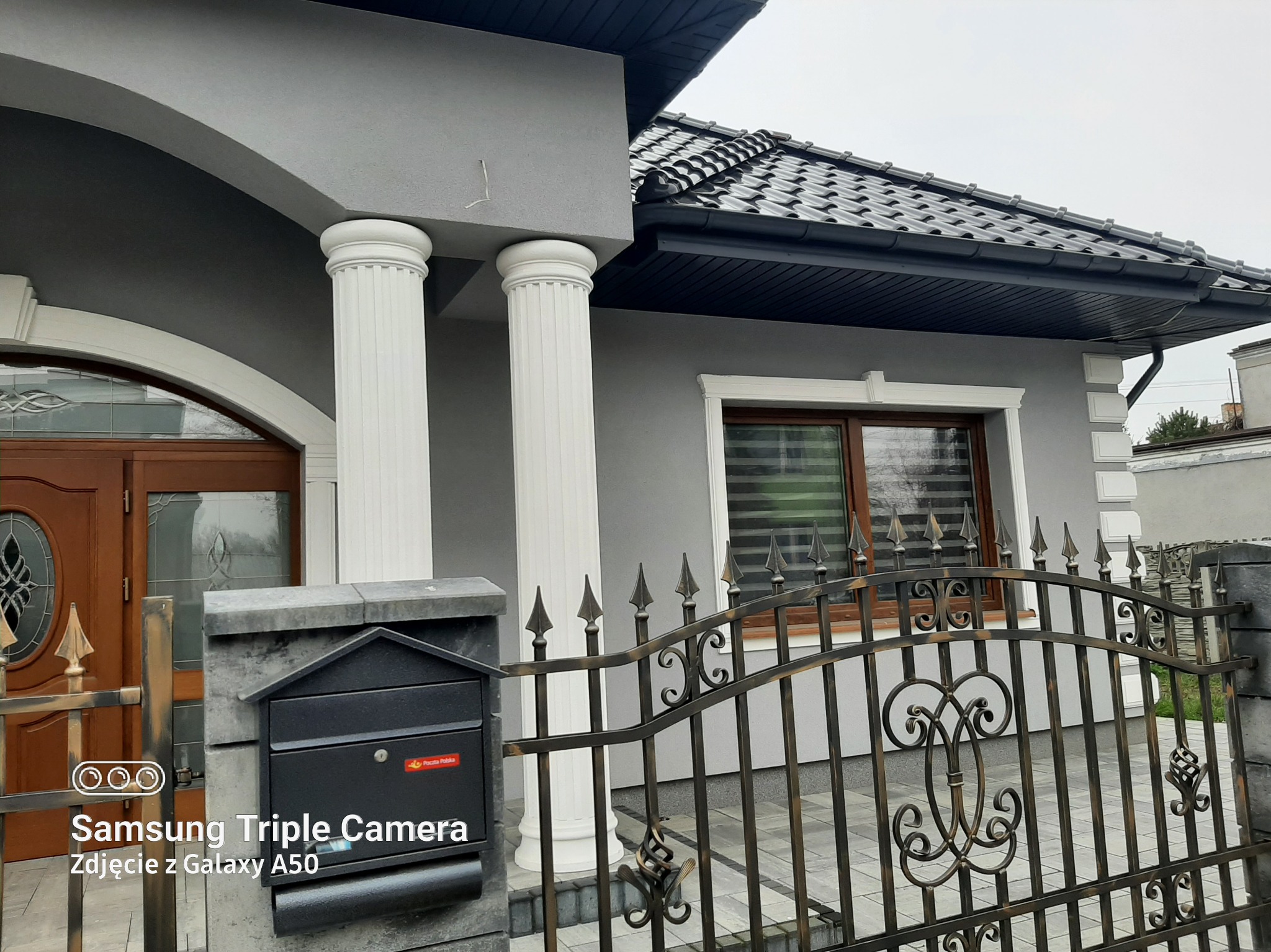 Elewacja domu w odcieniach szarości z białymi kolumnami i detalami. Widoczna brama z metaloplastyki i skrzynka pocztowa. Ujęcie z bliska, podkreślające estetykę wykończenia.