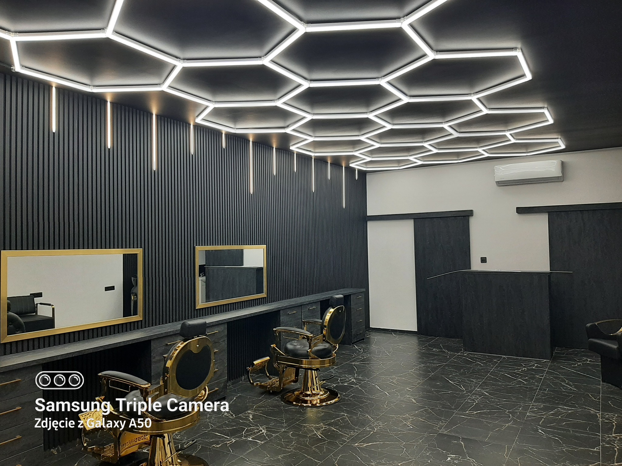 Nowoczesne wnętrze salonu fryzjerskiego z heksagonalnym oświetleniem sufitowym, czarnymi ścianami z pionowymi panelami i marmurową podłogą. Dwa złote fotele fryzjerskie.