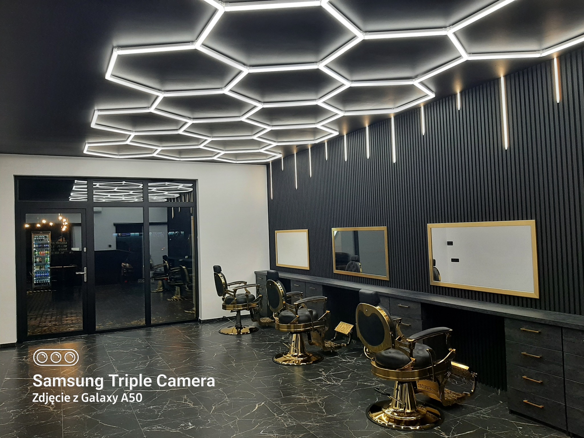 Nowoczesne wnętrze salonu fryzjerskiego z heksagonalnym oświetleniem sufitowym, czarnymi ścianami z pionowymi panelami i marmurową podłogą. Złote fotele fryzjerskie.
