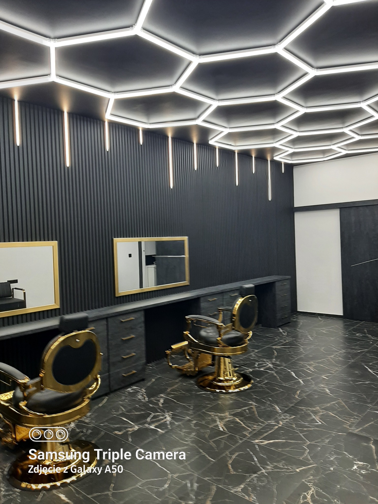 Nowoczesne wnętrze salonu fryzjerskiego: czarne ściany z lamelami, marmurowa podłoga, złote fotele i lustra, heksagonalne oświetlenie sufitowe.