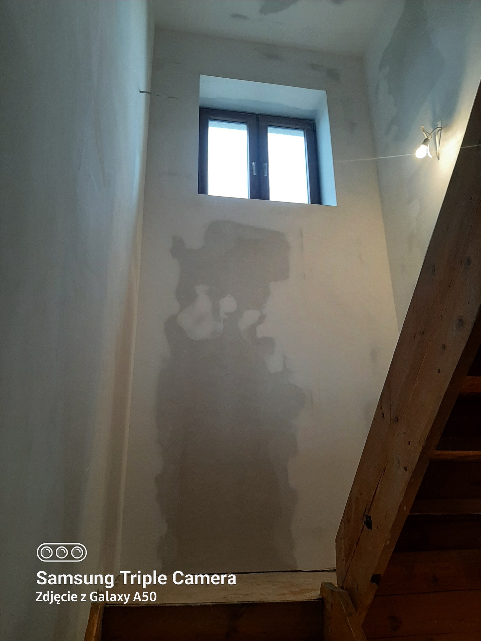 Ściana w trakcie gładzenia z widocznym oknem i fragmentem drewnianych schodów. Widoczne ślady po gładzi szpachlowej. Ujęcie z dołu, perspektywa podkreśla wysokość pomieszczenia.