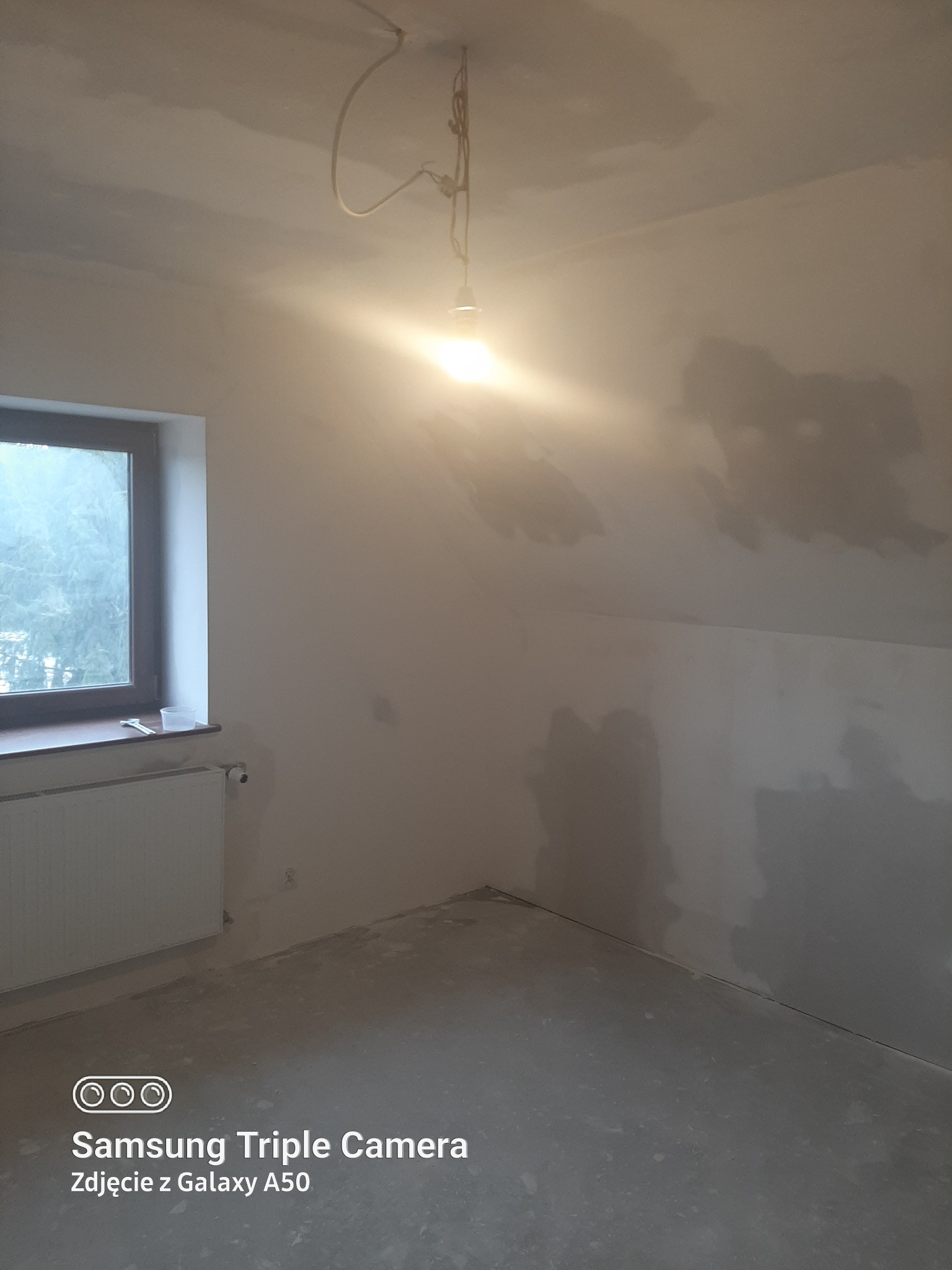 Pokój na poddaszu w trakcie prac wykończeniowych: ściany z widocznymi łatami gładzi, okno z parapetem, grzejnik. W tle widok za oknem. W pomieszczeniu wisi żarówka.