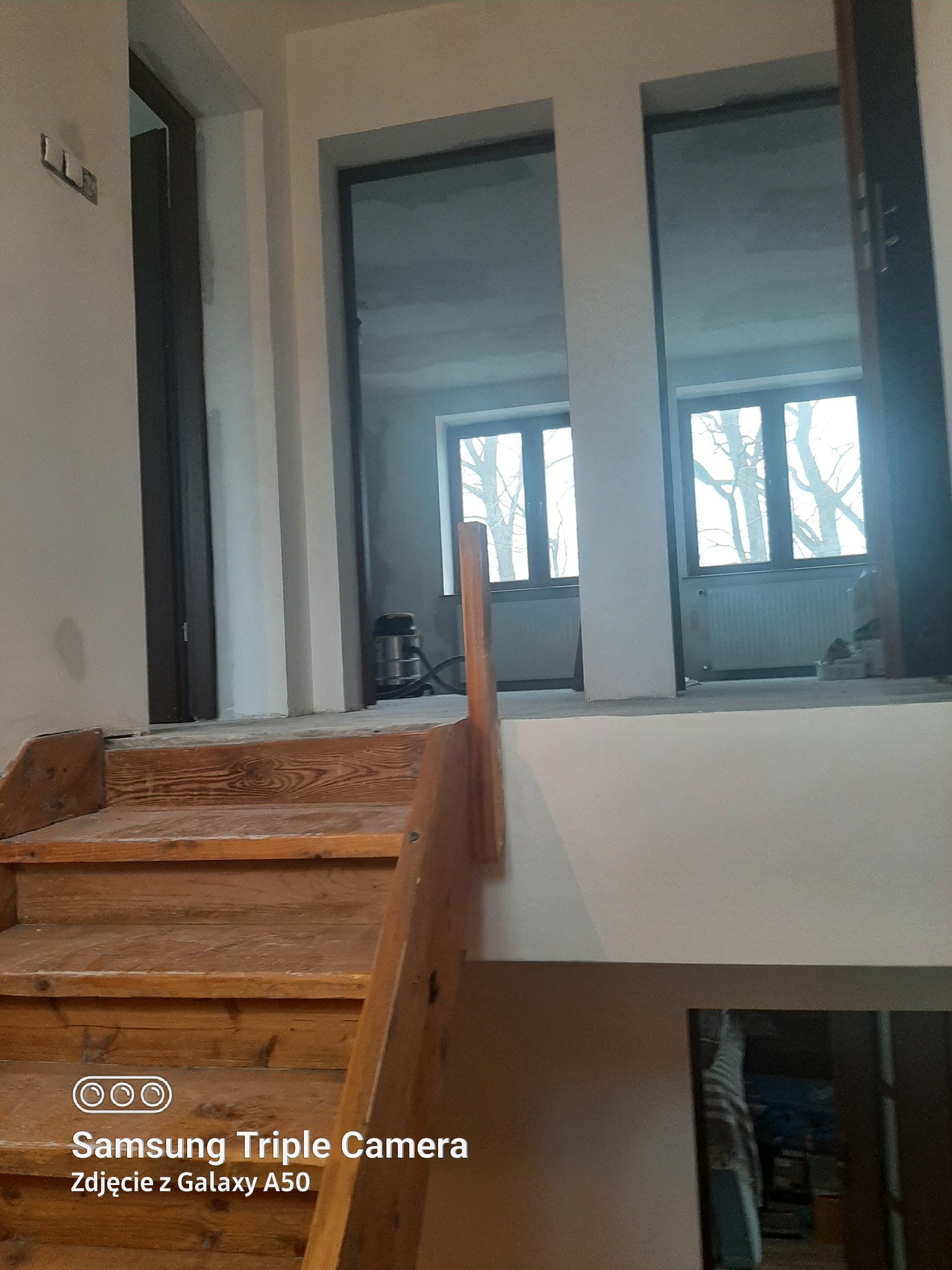 Wnętrze domu w trakcie remontu: drewniane schody prowadzą na antresolę z oknami. Widoczne ślady prac wykończeniowych, surowe ściany i narzędzia. Ujęcie z dołu.