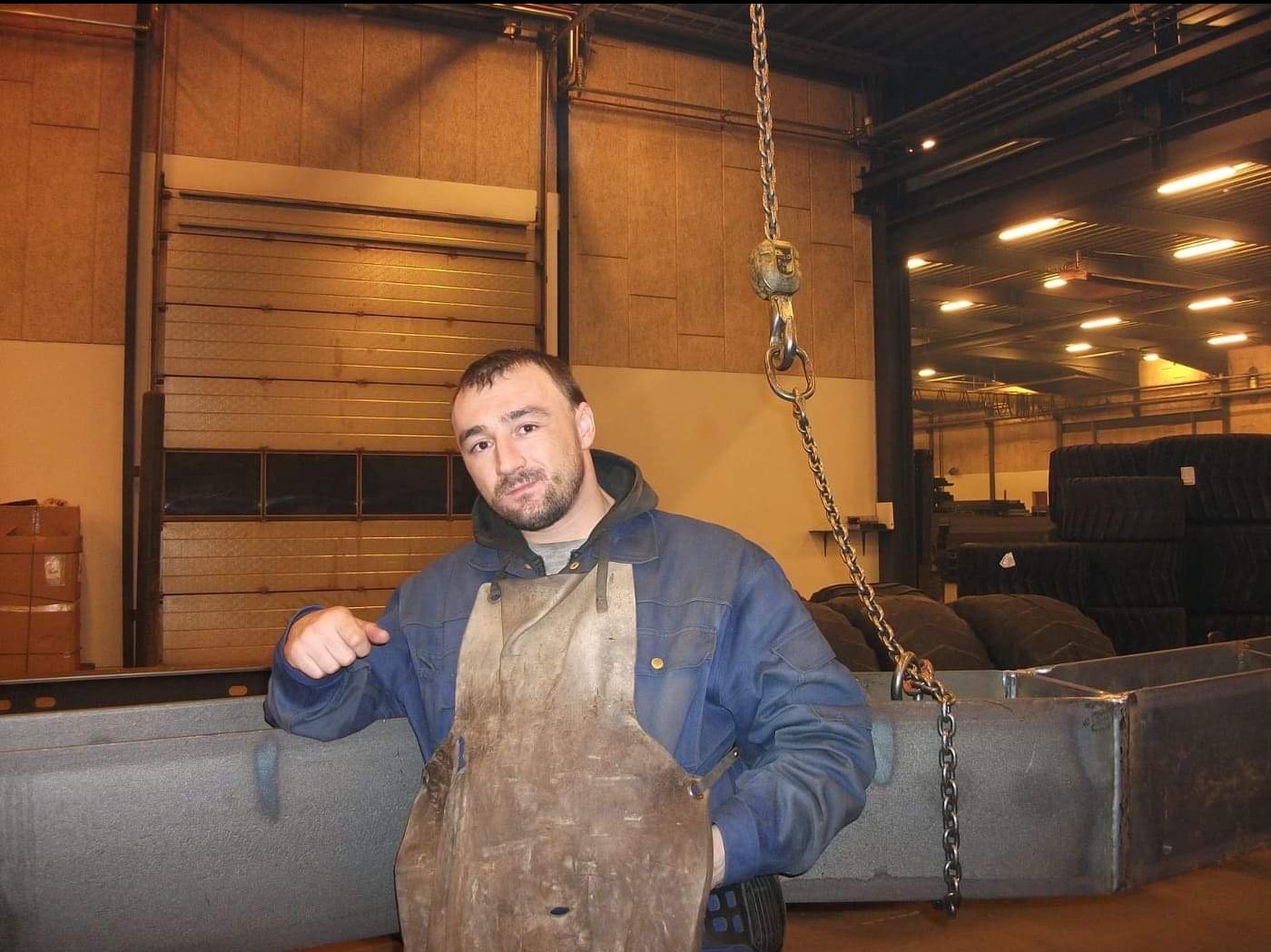 Portret ślusarza w skórzanym fartuchu, opierającego się o metalową konstrukcję podwieszoną na łańcuchu w hali produkcyjnej.