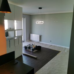 Łukasz Gałka Złota Rączka - Pokój w trakcie odświeżania: puszki z farbą i akcesoria malarskie na zabezpieczonej podłodze, ściany w odcieniach szarości, nowoczesna lampa sufitowa.