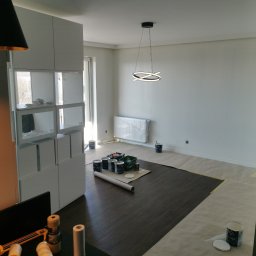 Łukasz Gałka Złota Rączka - Przygotowania do odświeżenia ścian w jasnym, przestronnym wnętrzu: podłoga zabezpieczona papierem, widoczne puszki farb i akcesoria malarskie, nowoczesna lampa sufitowa.