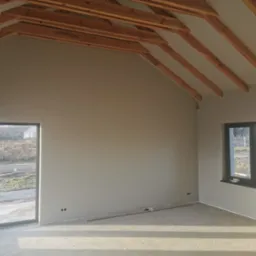 Surowe wnętrze domu w trakcie wykańczania: szare ściany po gładzi, betonowa podłoga, drewniana konstrukcja dachu, okna z widokiem na zewnątrz. Widoczne puszki elektryczne i listwa przypodłogowa.