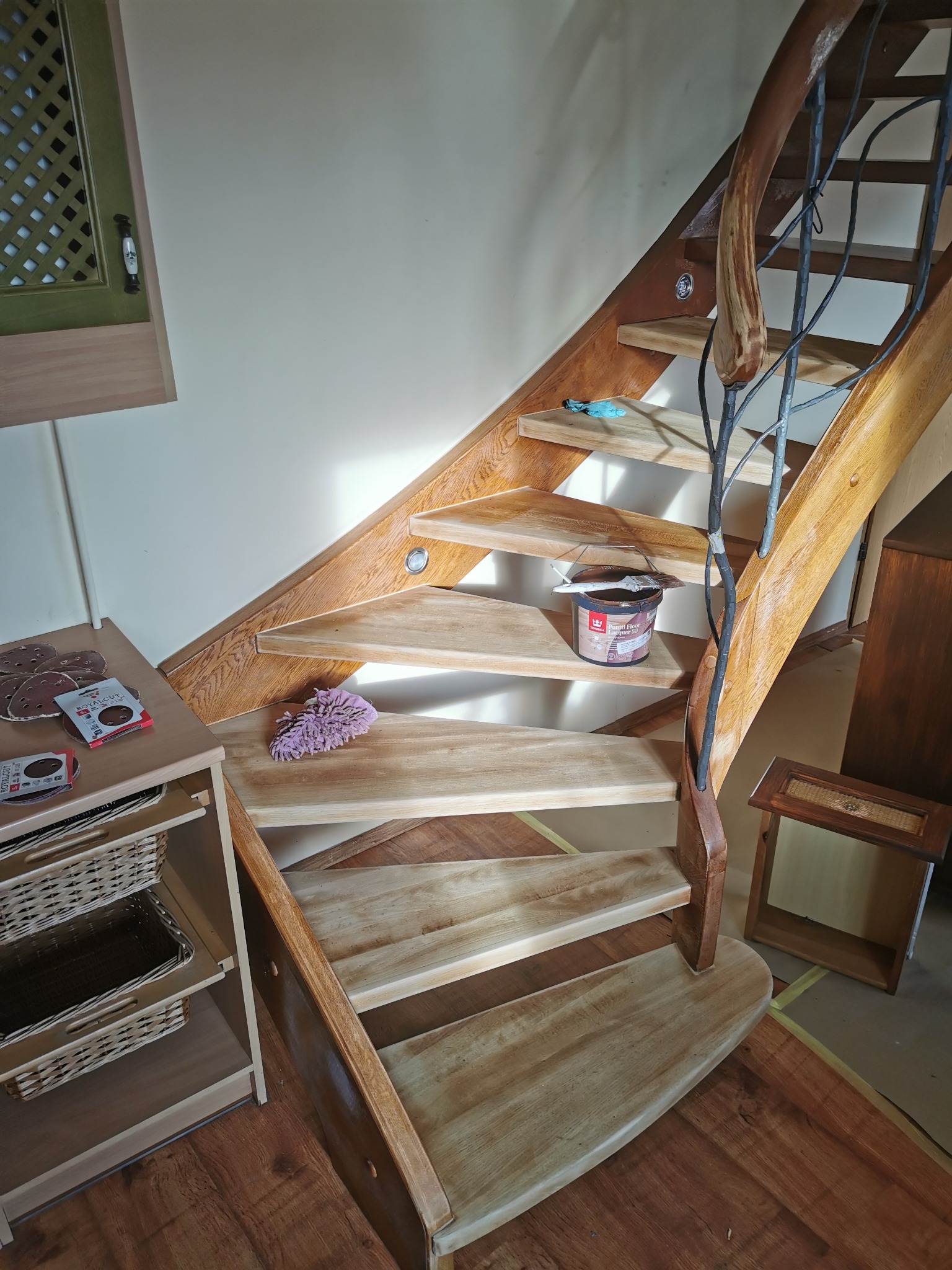 Drewniane schody z metalową balustradą w kształcie gałęzi, puszka z bejcą i pędzel na stopniu, widoczne materiały ścierne i ściereczka, etap renowacji schodów.