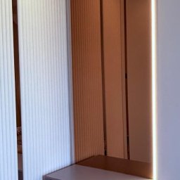 SZYMON PRUS DESIGN - Nowoczesna szafka z szufladą o ryflowanym froncie, wkomponowana w ścianę z dekoracyjnymi panelami. Oświetlenie LED podkreśla minimalistyczny design.