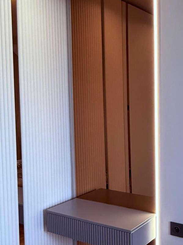 Nowoczesna szafka z szufladą o ryflowanym froncie, wkomponowana w ścianę z dekoracyjnymi panelami. Oświetlenie LED podkreśla minimalistyczny design.