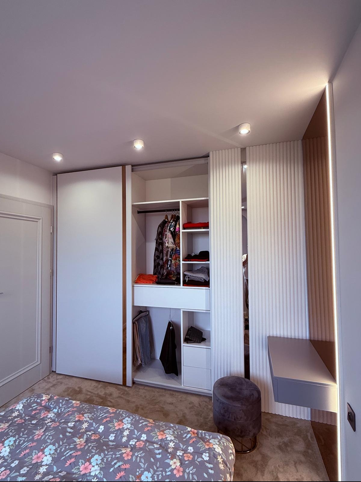 Nowoczesna szafa wnękowa z przesuwnymi drzwiami i oświetleniem LED, widoczna z perspektywy łóżka w sypialni. Minimalistyczny design, białe fronty i drewniane akcenty.